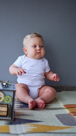 cute serious baby wearing white bodysuit sits at the wall indoors. child looks up and then pays attention to his toy. vertical video.