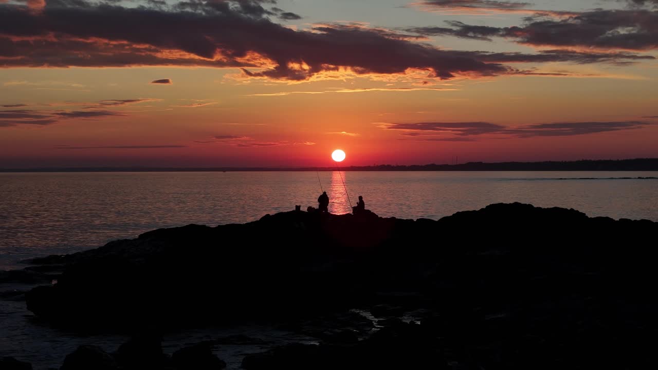 Drone orbits fishermen on rocks during a magical sunset at the coast of Uruguay. The sun glows before setting, reflecting over the water. Sky and clouds painted in warm colors