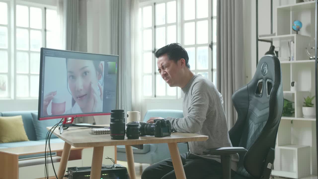 Asian Cameraman In Long Sleeved T-Shirt And Black Pants Having Backache While Using Desktop Computer For Editing Photo At Home.
