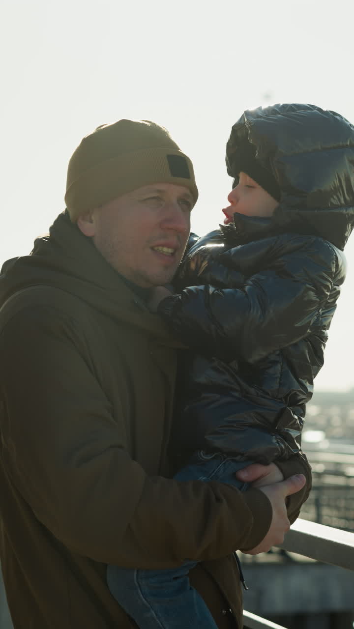 A father stands close to a metal railing, carrying his son who gently places his hand on the father s shoulder, gazing out over a cityscape in the distance