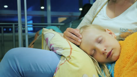 a woman is stroking a girl sleeping on her lap sitting in the terminal of the airport flight delay c