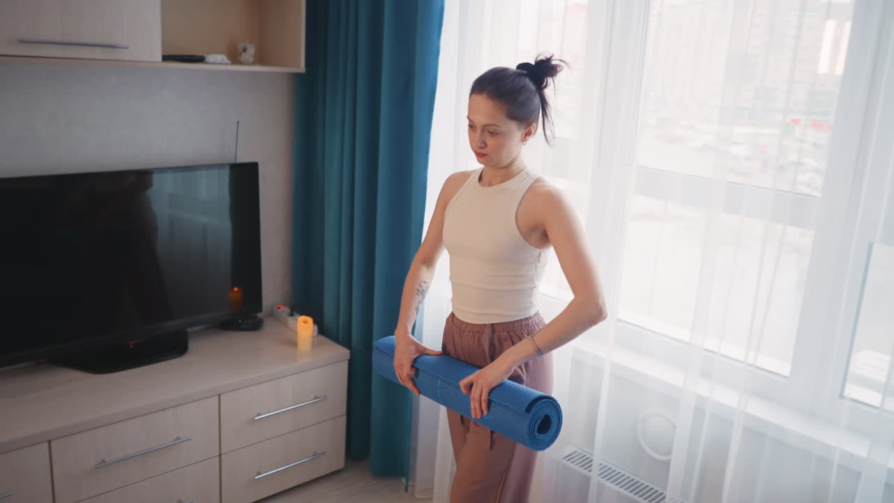 Giovane donna che srotola un tappetino da yoga blu sul pavimento in legno, candela accesa accanto al mobile TV, luce mattutina soffusa che filtra dalle tende, preparazione a piedi nudi per una rilassante routine di yoga, atmosfera domestica delicata