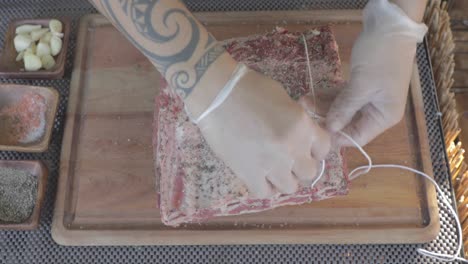 close up top view of chef tied piece of steak with twine on wooden board of restaurant kitchen.