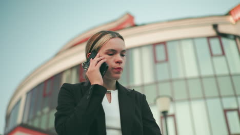fed up lady in professional outfit speaks on phone with tired expression in urban setting, modern building slightly blur in background