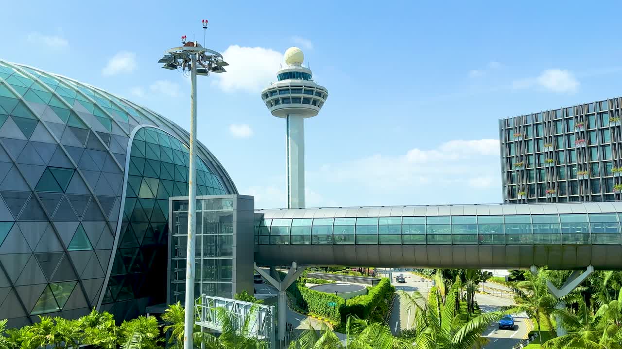 A smooth daytime camera pan reveals a glass-domed airport terminal, lush greenery, and a prominent air traffic control tower under clear blue skies
