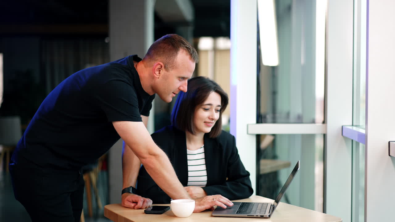 Positive young Caucasian lady sits at the desk. Male colleague leaned over her to the laptop. Cooperation at office work.