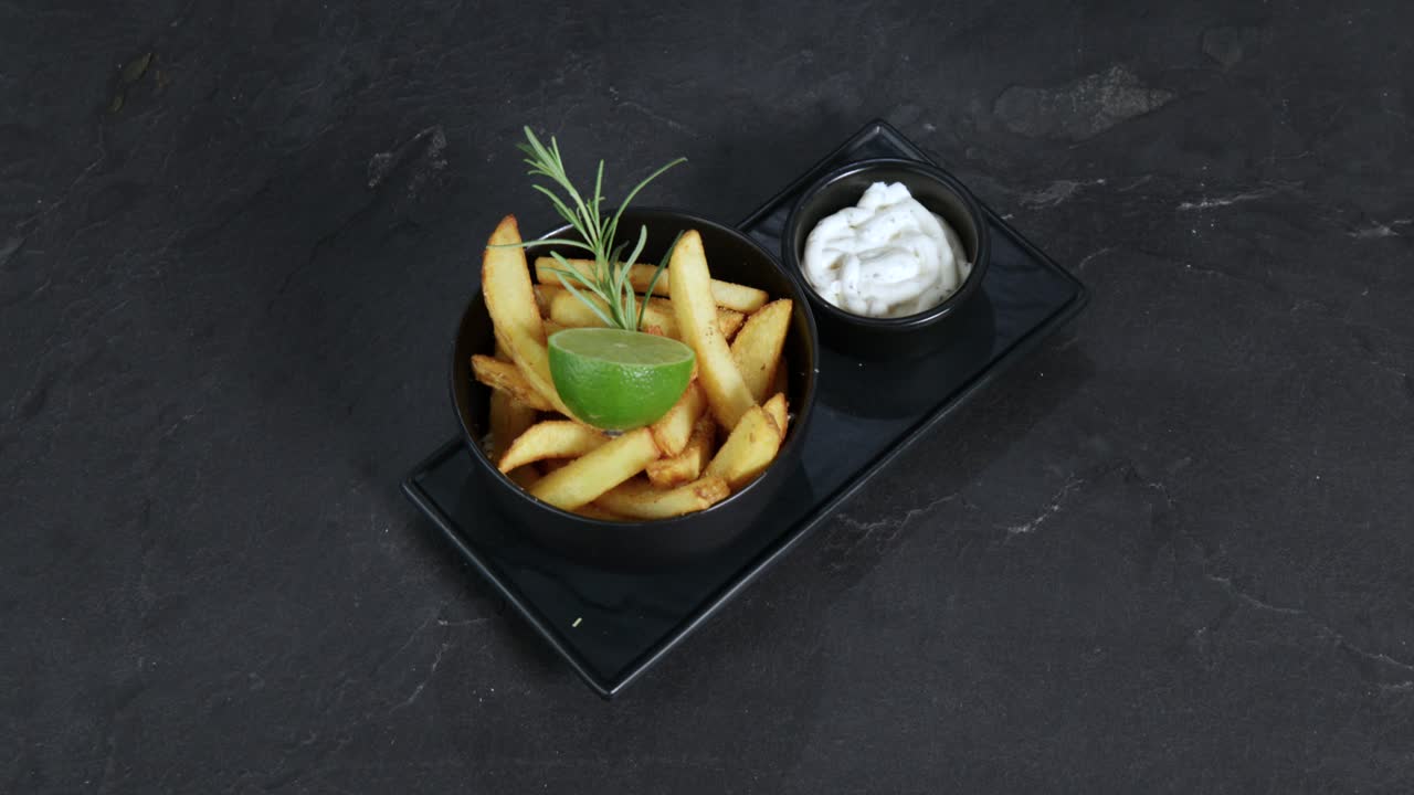 French fries garnished with rosemary and lime next to a mug of mayonnaise are presented on a rotating plate