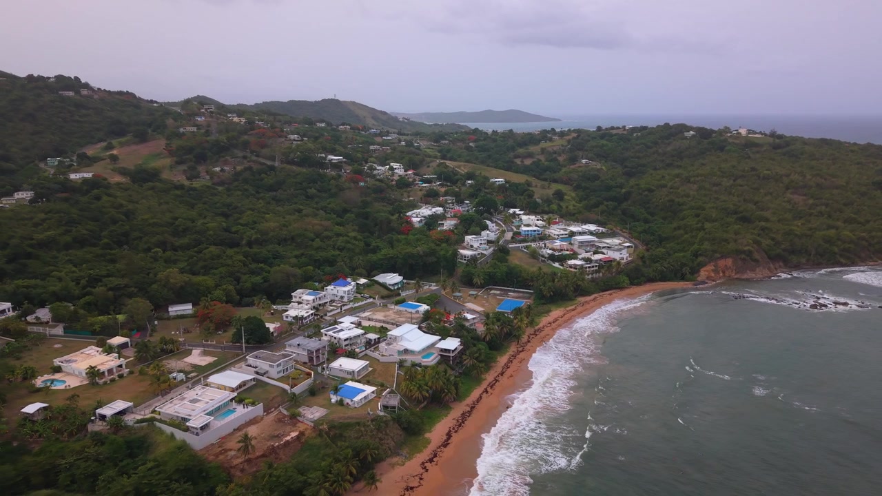 Pemandangan udara dari atas garis pantai tropis berpasir dan desa pesisir yang terletak di antara perbukitan hijau subur