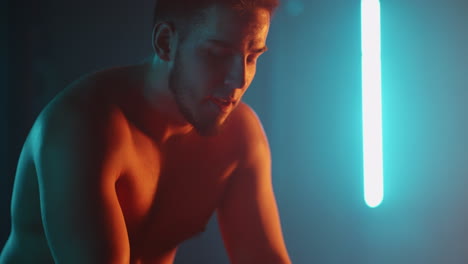 a tired athlete is resting in breathing heavily while sitting on a barbell in neon light after performing an exercise in slow motion. recuperate and prepare for a new breakthrough