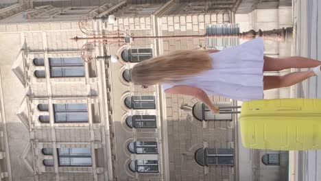 a young beautiful girl with a yellow suitcase is walking on a city street from behind. back view of a girl traveler walking and pulling a suitcase