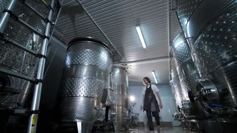 a happy successful farmer or winemaker is walking in the middle of wine tanks and checking a fermentation grade of high quality wine production in a modern winemaking facility or winery.