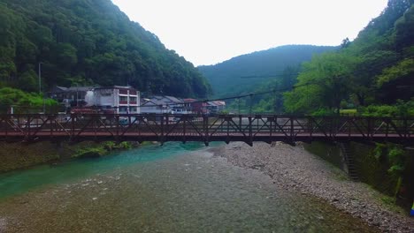 Drohne-Steigt-über-Den-Fluss-Und-Enthüllt-Nach-Dem-Überqueren-Einer-Brücke-Ein-Japanisches-Dorf-In-Einem-Tal