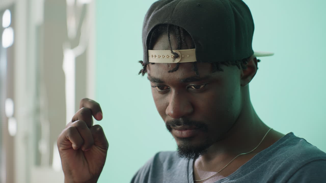 Close up of calm young man wearing dark cap backward and casual shirt, looking downward with thoughtful expression, hand raised near face, standing indoors against turquoise background