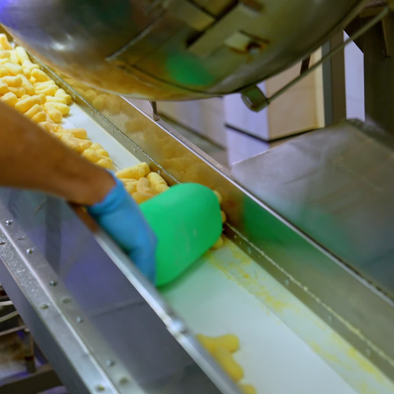 Factory worker takes corn sticks out of a metal tank. Crispy puff corns are put on the conveyor belt for the next stage of production
