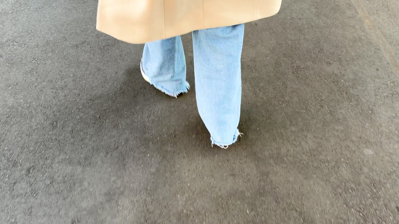 Una persona in jeans blu e un trench cammina su un marciapiede asfaltato all'aperto.