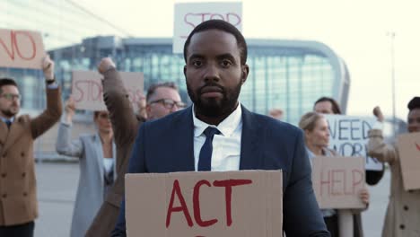 camera zoom out on an african american man holding act now" signboard in a protest with multiethnic business colleagues in the street"