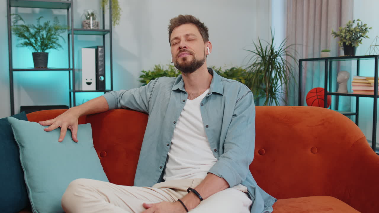 무선 헤드폰을 착용한 행복한 편안한 남자가 집 소파에서 좋아하는 록 앤 롤 음악을 듣고 춤을 추고 있습니다.