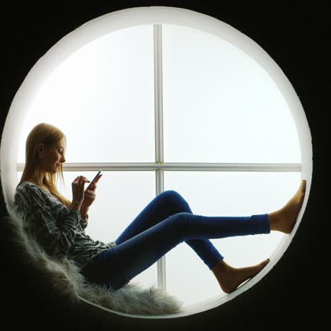 niña sentada en una ventana redonda usando un teléfono inteligente