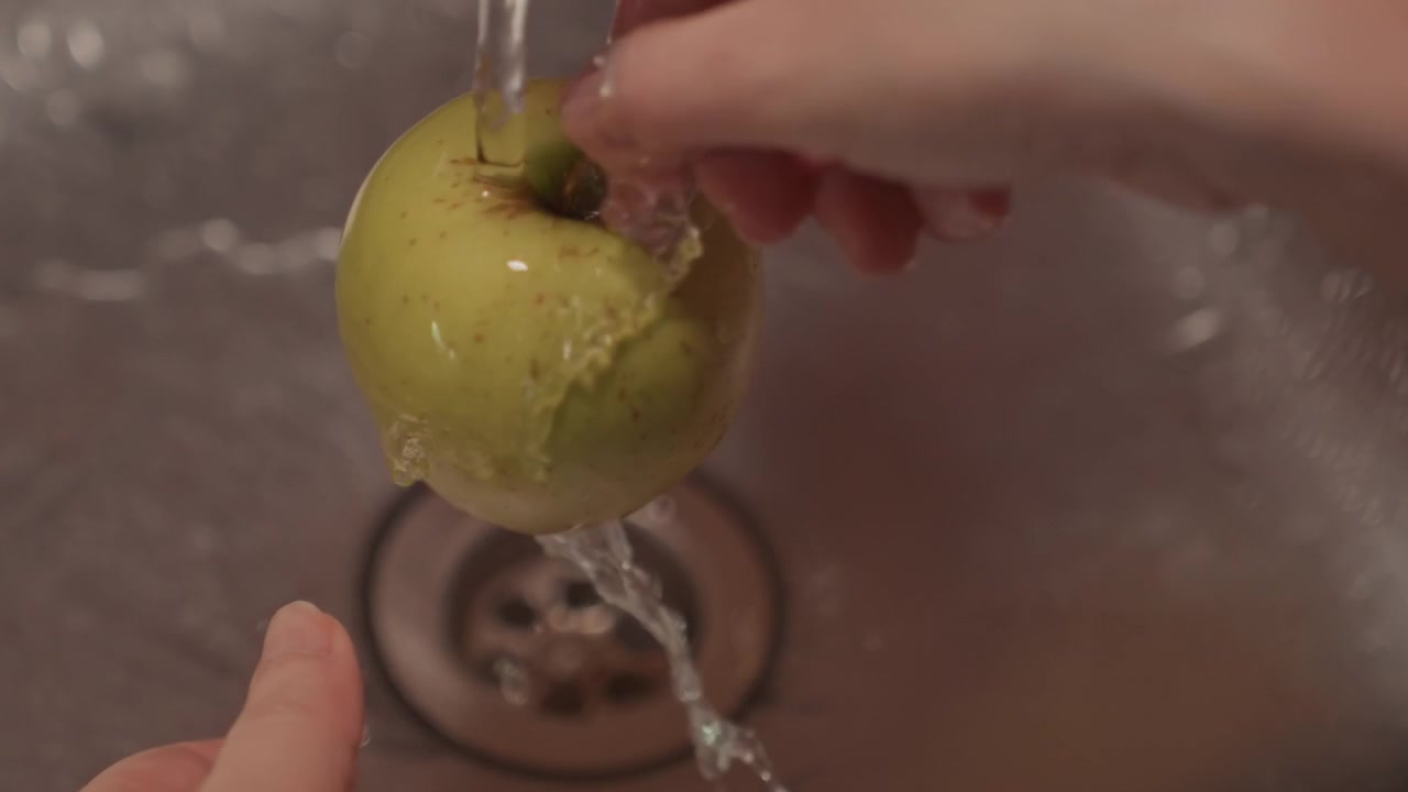 Primo piano di mani che lavano una mela verde sotto acqua corrente nel lavello. Il frutto viene ruotato e sciacquato accuratamente con un getto d'acqua limpida. Lo scarico del lavello è visibile per tutta la sequenza.