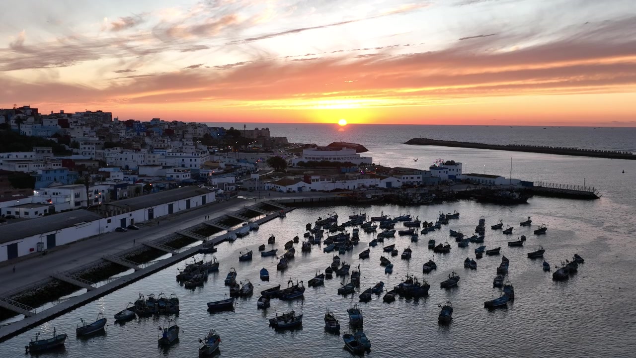 Cảnh quay chậm từ trên cao hoàng hôn trên cảng cá Asilah thuộc bờ biển Đại Tây Dương của Maroc, với hàng chục thuyền đánh cá nhỏ, mặt nước tĩnh lặng, bầu trời tối đầy màu sắc và thành phố ven biển lịch sử ở phía sau