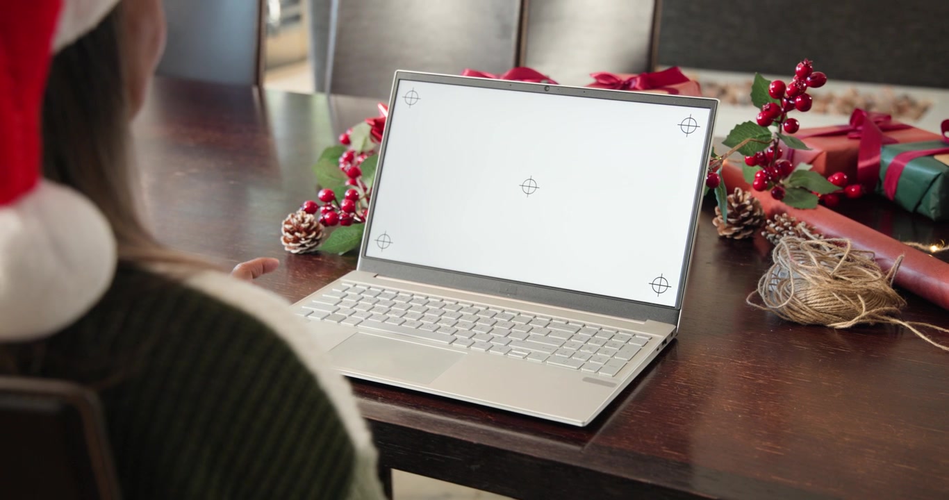 Donna con cappello di Babbo Natale alza la mano verso il laptop all'inizio di una chiamata, salutando durante una chat festiva al tavolo.