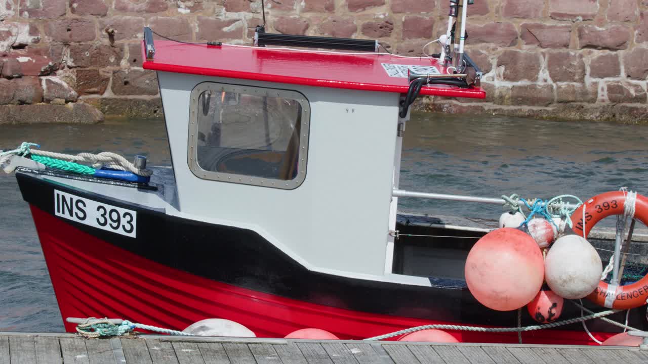 Stationary red fishing boat with marine equipment beside stone harbor wall, daylight, minimal camera movement