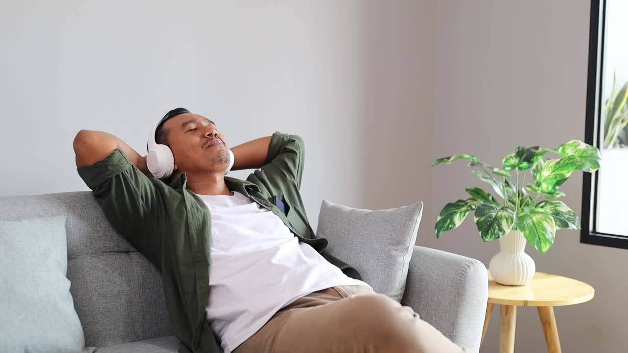 Uomo asiatico che ascolta musica in cuffie wireless, seduto sul divano. Relax del fine settimana.