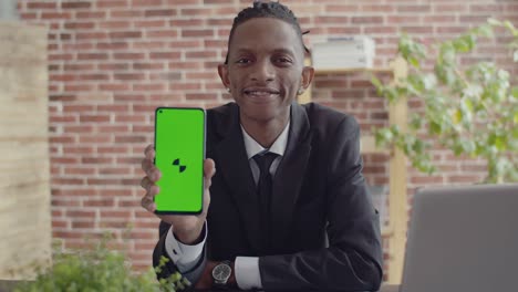 black man businessman in an official suit sits at a desk in office and smiles shows the green screen of phone and looks into the camera