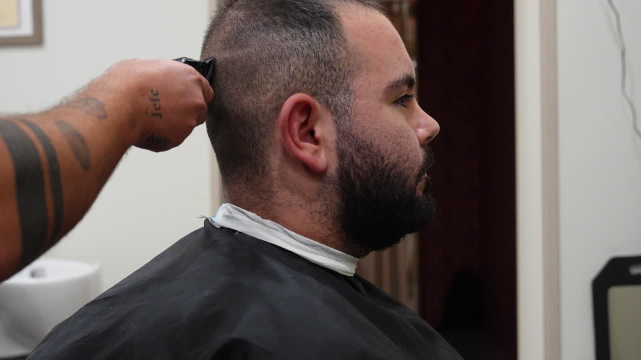 Profile shot of a man seated in barbershop while a tattooed hairdresser cuts his hair with a trimmer