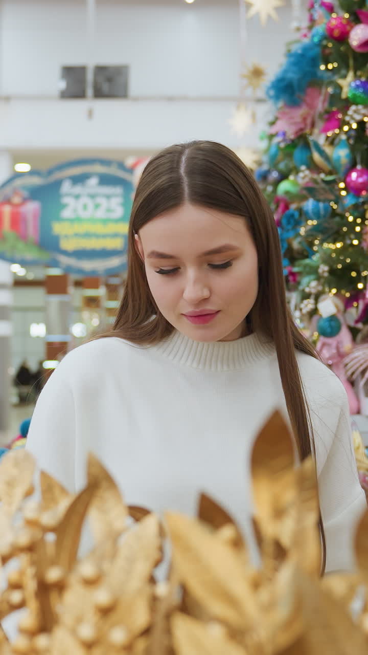Woman in decor store holding and admiring a brown decorative ornament with a smile, surrounded by vibrant holiday decorations and other shoppers in the background