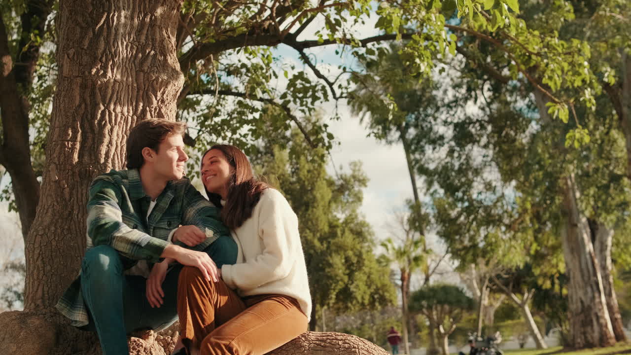 Happy young lovers man and woman relaxing sitting in park under tree