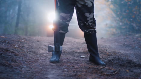 piernas con botas de un hombre que sostiene un hacha, los faros de un auto brillan detrás de una espeluznante escena del crimen