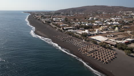 Aerial-View-of-Parasols-and-Waterfront-Buildings-on-Black-Sand-Beach,-Santorini-Island,-Greece