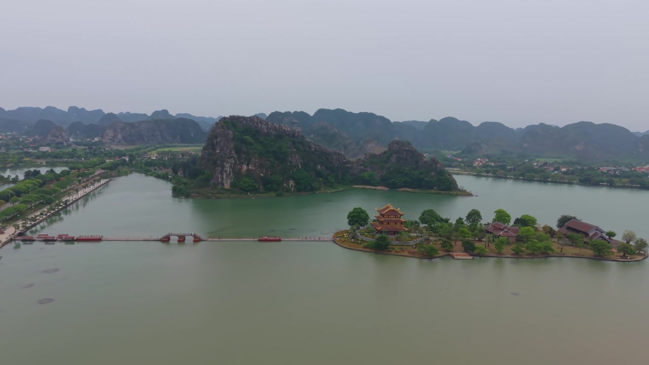 Cảnh quay drone kéo lùi tiết lộ chùa Bái Đính trên một hòn đảo được bao quanh bởi làn nước tĩnh lặng và phong cảnh núi đá vôi ở Ninh Bình, Việt Nam