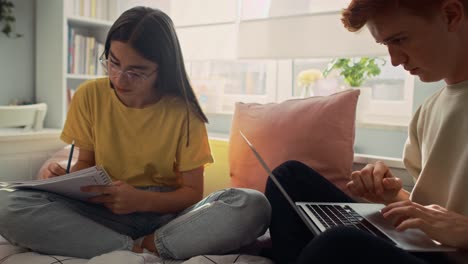 dos adolescentes caucásicos sentados en la cama y aprendiendo de libros y computadoras portátiles