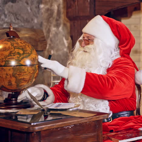 santa with a globe. mature man in red costume of santa sitting at the table in the room and looking at the globe. christmas time.