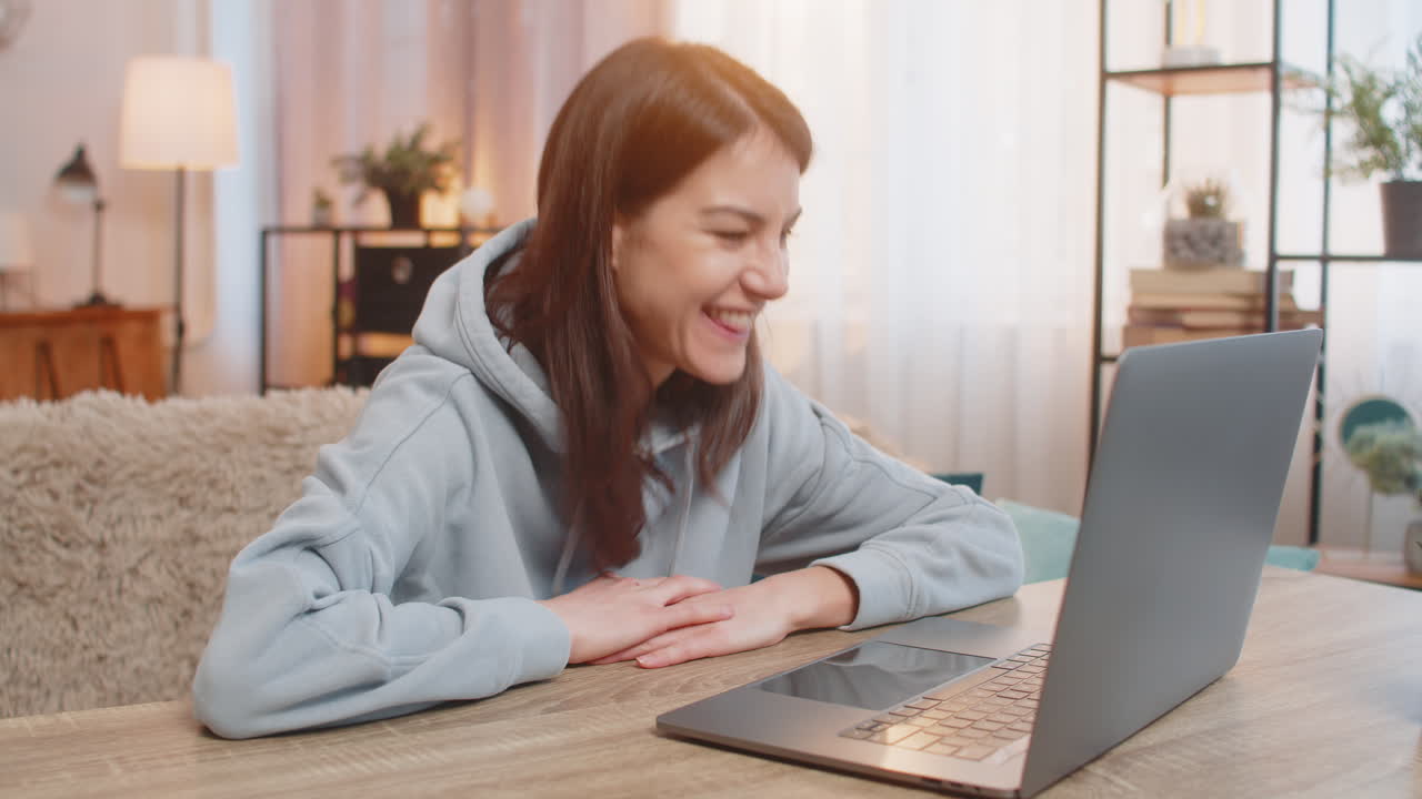 Happy woman sitting on sofa at table using laptop celebrating online victory or good news at home