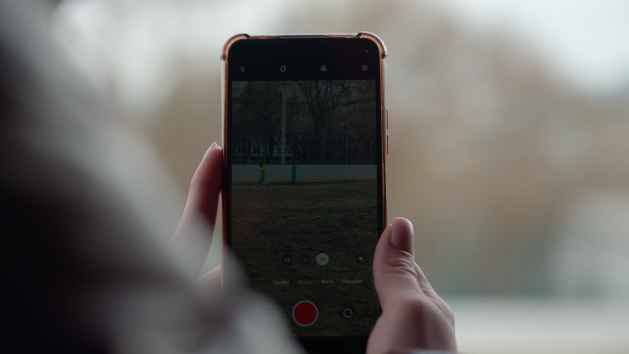 Grabación con el móvil por encima del hombro de un niño en un parque, silueta difuminada en primer plano, tonos otoñales y luz tenue, composición espontánea que captura el movimiento y la práctica desde el punto de vista de un padre o entrenador