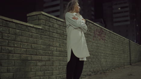 confident woman in white coat stands outdoors at night by brick wall with arms folded, appearing deep in thought. urban backdrop includes high-rise buildings and soft ambient street lighting