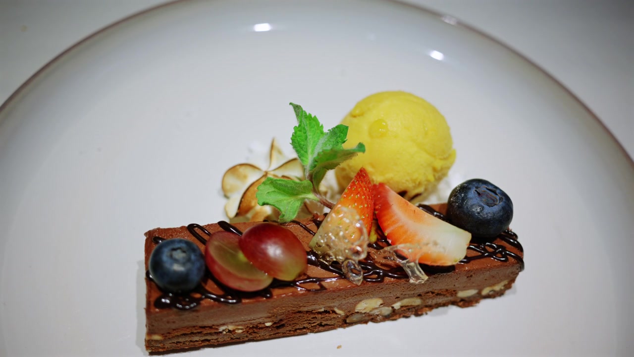 Nærbilde av en elegant dessert servert på en restaurant, med bær, krem, mynte, sjokolade og en kule gul sorbet
