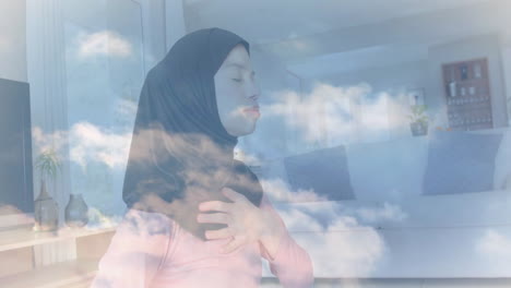 animation of clouds over biracial woman in hijab practicing yoga