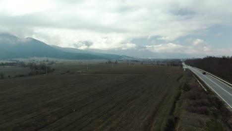 Drone-shot-over-country-road-with-moving-cars-and-mountains-view-with-cloudy-sky