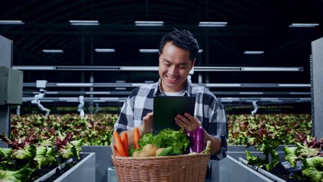 asian male farmer with vegetable basket smiling while using tablet in smart farm