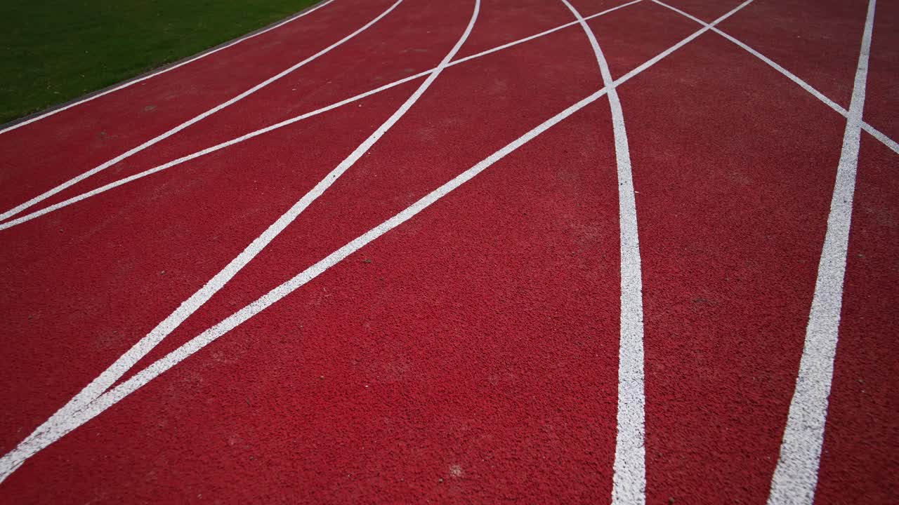 Following dark red running track crossed by the white lines. Green grass field and paved path on both sides of the track. Close up.