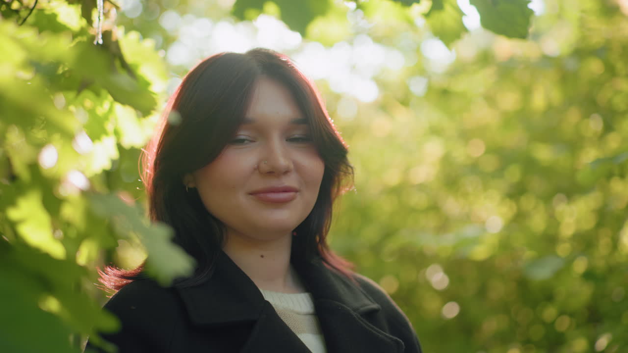 Portrait of nature lover standing in arboretum, warm sun beam on face, soft smile bright, hair glowing with light, leafy bokeh around, calm mood, relaxed pause during quiet autumn stroll