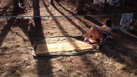 young woman resting reading book in camp in wild pine forest at sunset