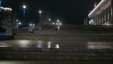 nighttime view of two people walking up a wide, illuminated urban staircase, with tranquil ambiance of the city at night, and street lamps with architectural details