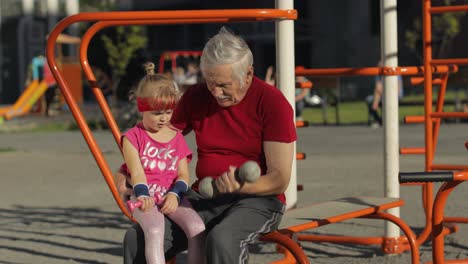 Granddaughter-and-grandfather-doing-fitness-exercises-with-dumbbells.-Senior-man-with-child-kid-girl