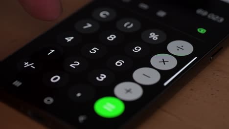 a close-up of a finger pressing a button on a smartphone calculator app. the bright numeric keys and green equals button stand out against the dark background, emphasizing modern digital calculation