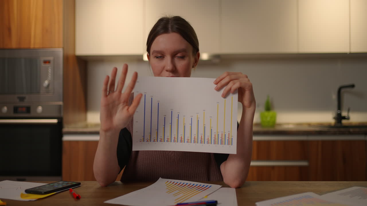Home office Looking at the camera a young woman shows a graph to the camera and gestures shows and explains the data values and explains the company's analytics. Course paper